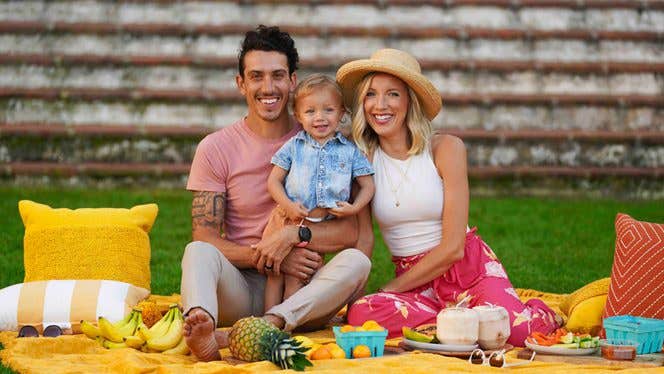 Erin and Dusty Success Story A young couple sit on a picnic blanket with their toddler between them, all smiling. Fruit including pineapple and coconut is spread out on the picnic blanket before them.