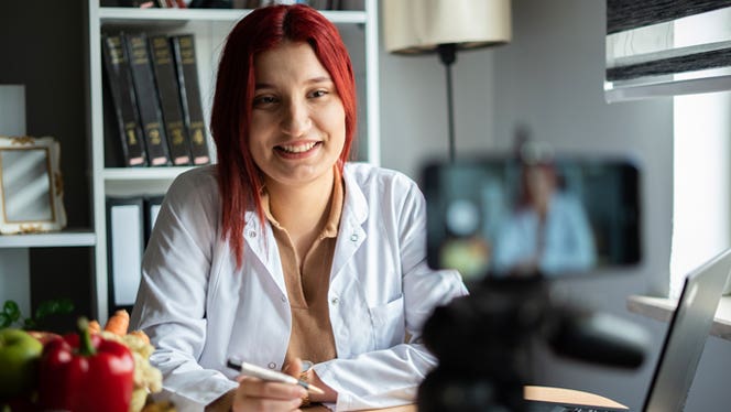 plant-based-telehealth-appointment A doctor sits with fruits and vegetables on her desk and a laptop and phone setup for a telehealth appointment